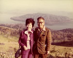 An old snapshot of a middle-aged Japanese couple on vacation. They stand shoulder to shoulder, smiling on a sloping hillside. Below and behind them, a body of water with a small island.